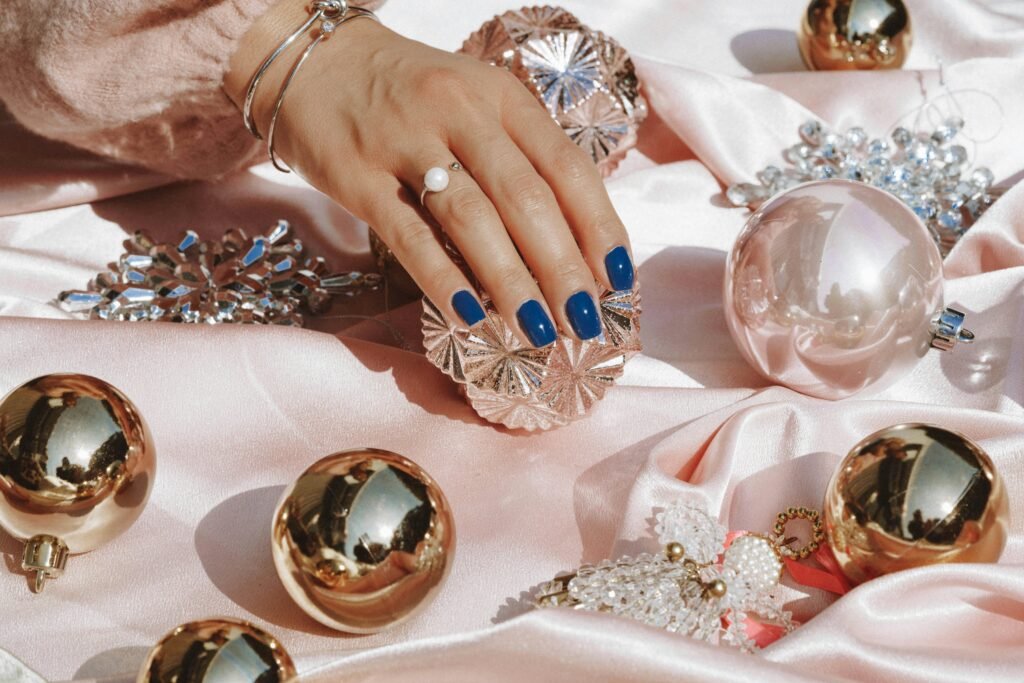 Elegante mano con uñas azules descansando sobre satén rosa rodeada de opulentos adornos y joyas.